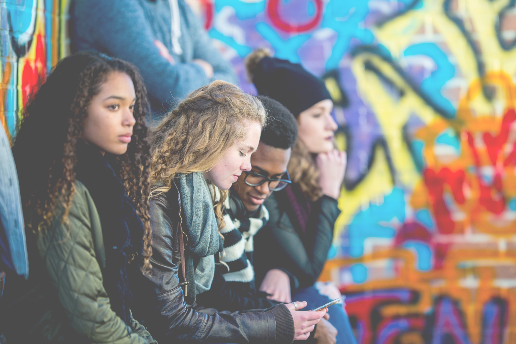 A group of youth sit along a bright colored, graffiti style mural.