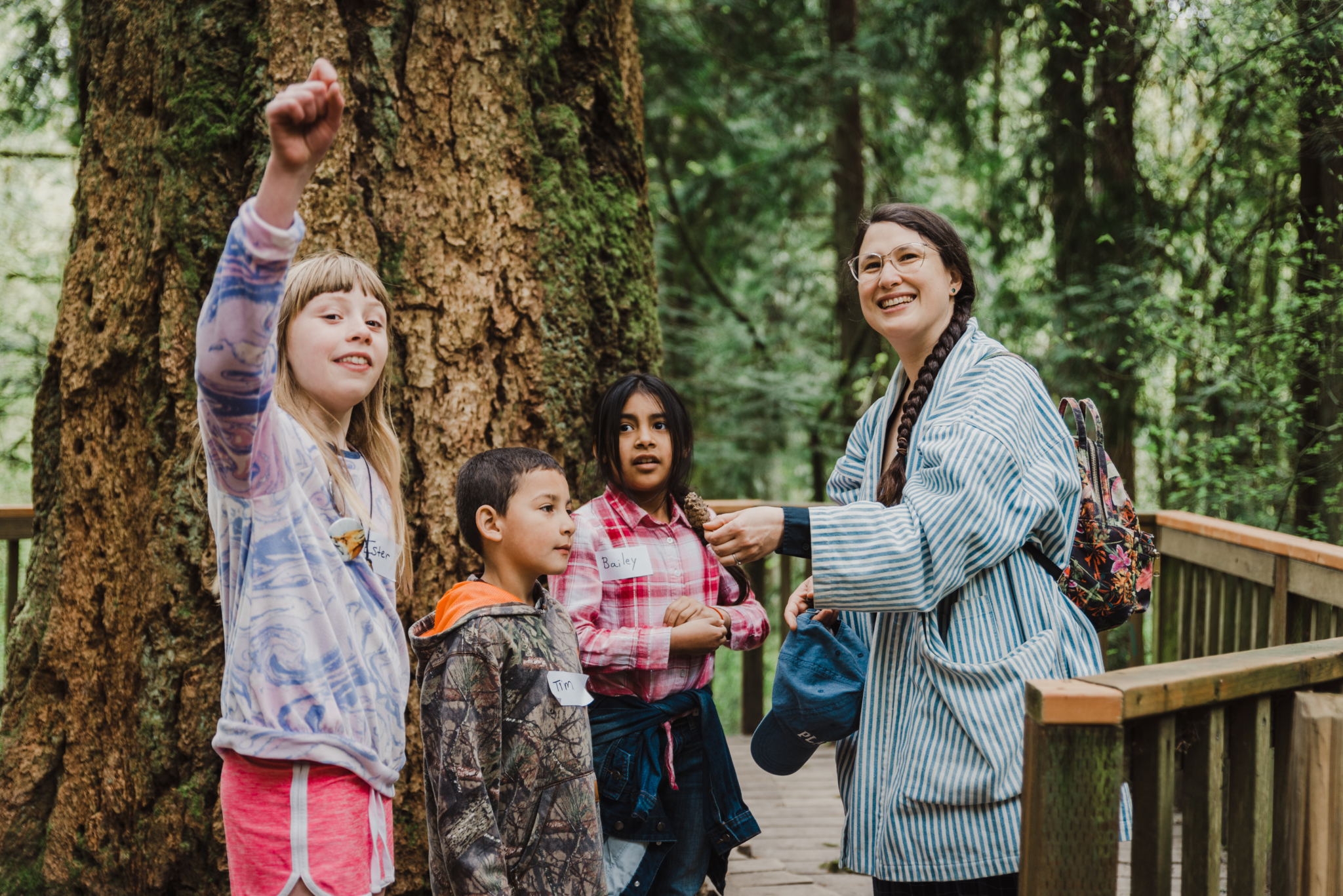 A smiling adult leads a group of kids on a forest adventure.