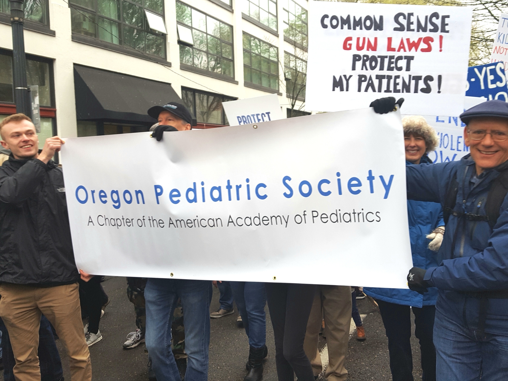 A group of people march holding a banner that says "Oregon Pediatric Society. A chapter of the American Academy of Pediatrics." Behind these people are additional marchers holding signs about "common sense gun laws! Protect my patients!"